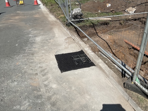 A photo of a new drain cover added, with fresh black tarmac.