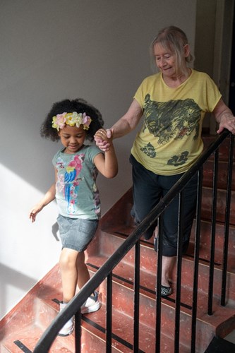 A senior woman and a girl aged about six are walking down a staircase holding hands. They are dressed in summer clothes and sunshine comes through a window