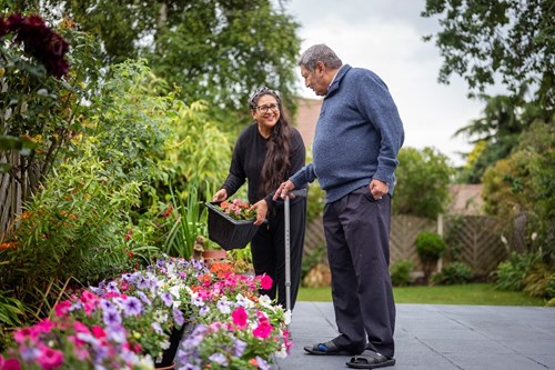 A man using a walking stick is talking to a woman who holds a tub of flowers. They are outside enjoying the flowers and plants in a beautiful garden border