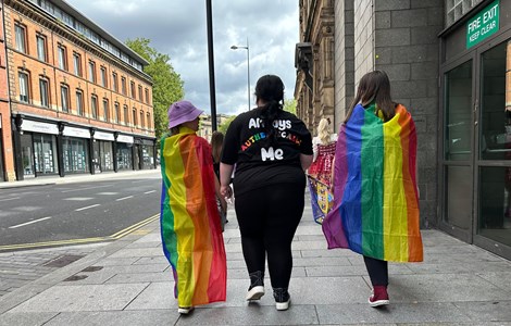 People walking away from the camera. Two are draped in rainbow flags, with the centre person wearing a shirt that says "always authentically me".
