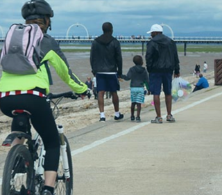 An image of a cyclist and pedestrians walking down alongside the beach, on a marked path. Southport pier is in the background. 