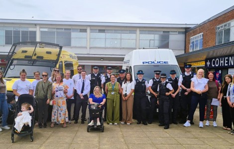 A large group of community members and police officers pose for a group photo in an outdoor shopping precinct. The group stands in front of two police vehicles, including a yellow police van on the left and a white police van on the right. Several adults are accompanied by young children, including one child seated in a pushchair and another partially visible beside it. One person is using a mobility scooter at the front of the group. 