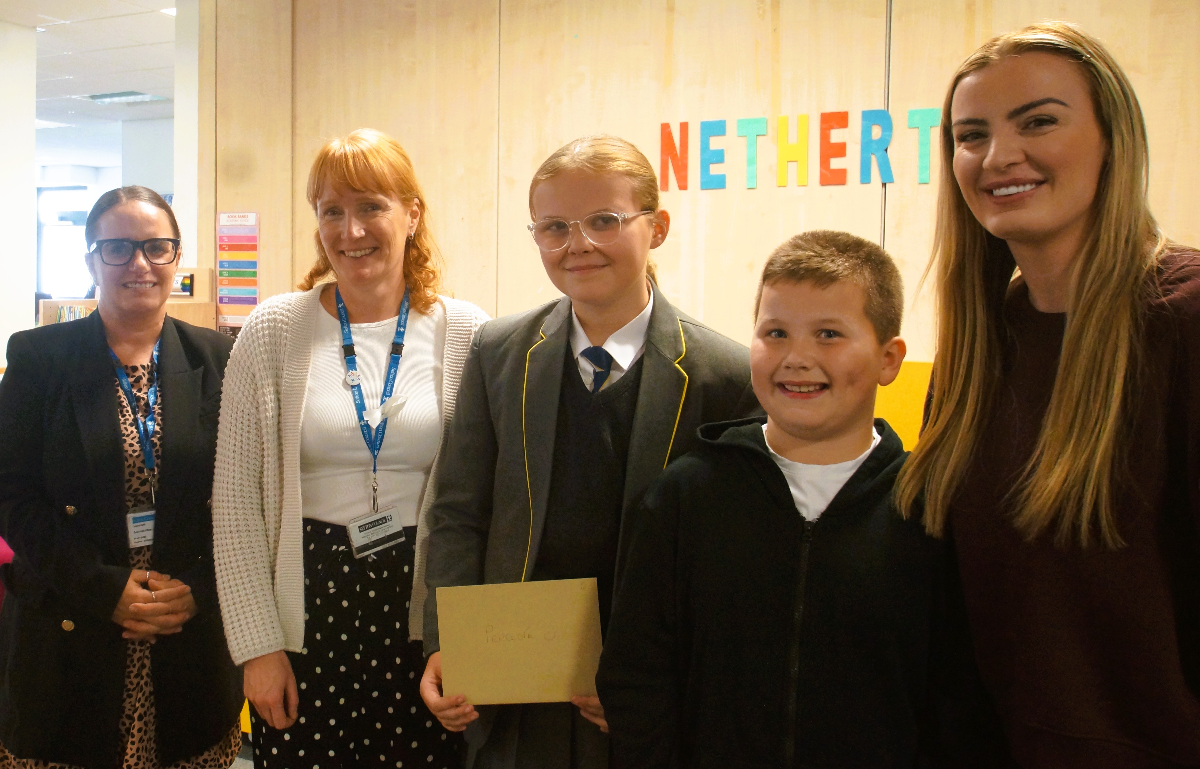 Five people stand together. One person holds a tan envelope with "Penelope U" written on it. Colourful letters spelling Netherton