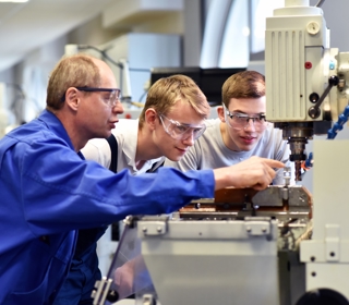 An instructor in a blue lab coat and safety glasses points to a machine while two students in safety glasses look on.