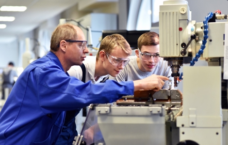 An instructor in a blue lab coat and safety glasses points to a machine while two students in safety glasses look on.