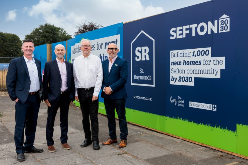Four men stand in front of a blue construction barrier that reads "SEFTON 2030 Building 1,000 new homes for the Sefton community by 2030."